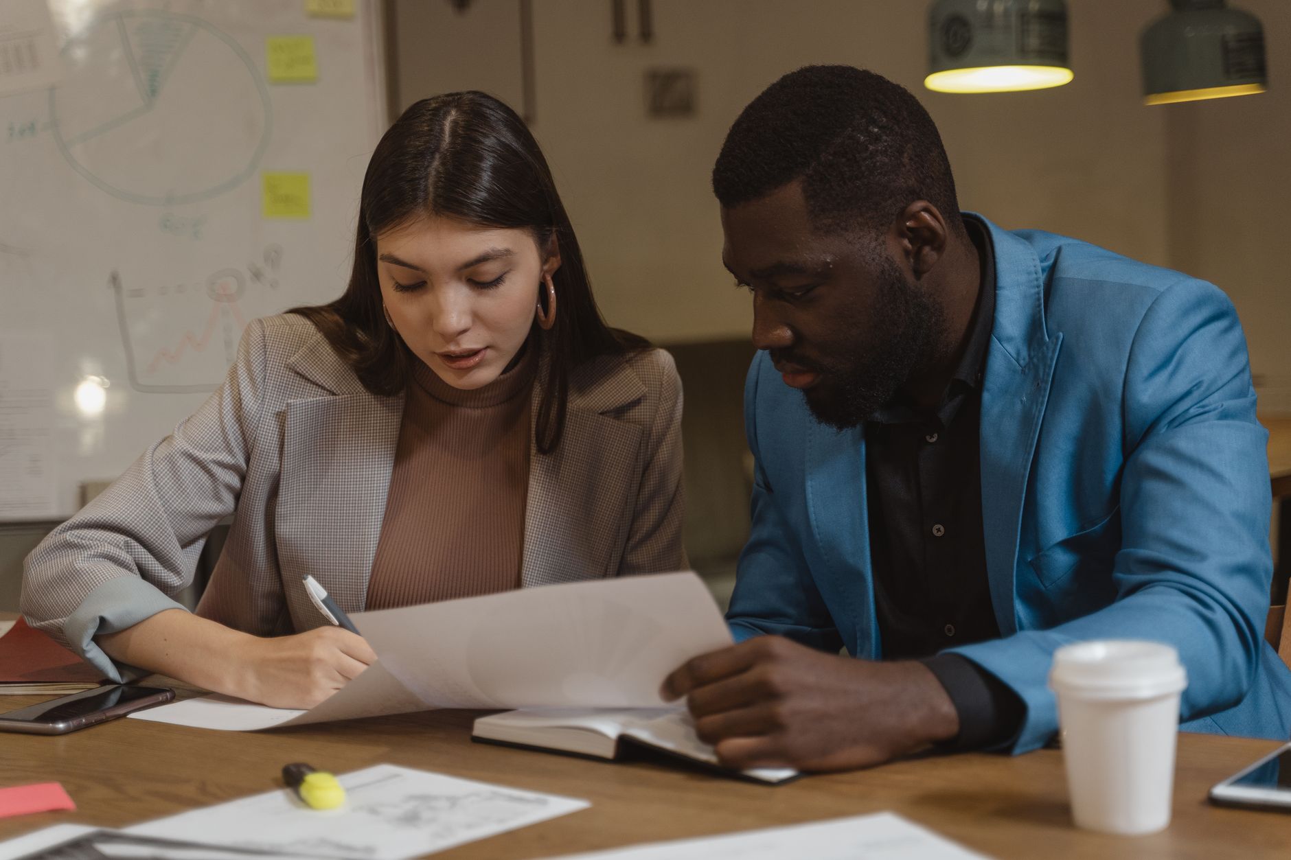 Deux collègues examinent ensemble des documents papier à une table de bureau, devant un tableau blanc.