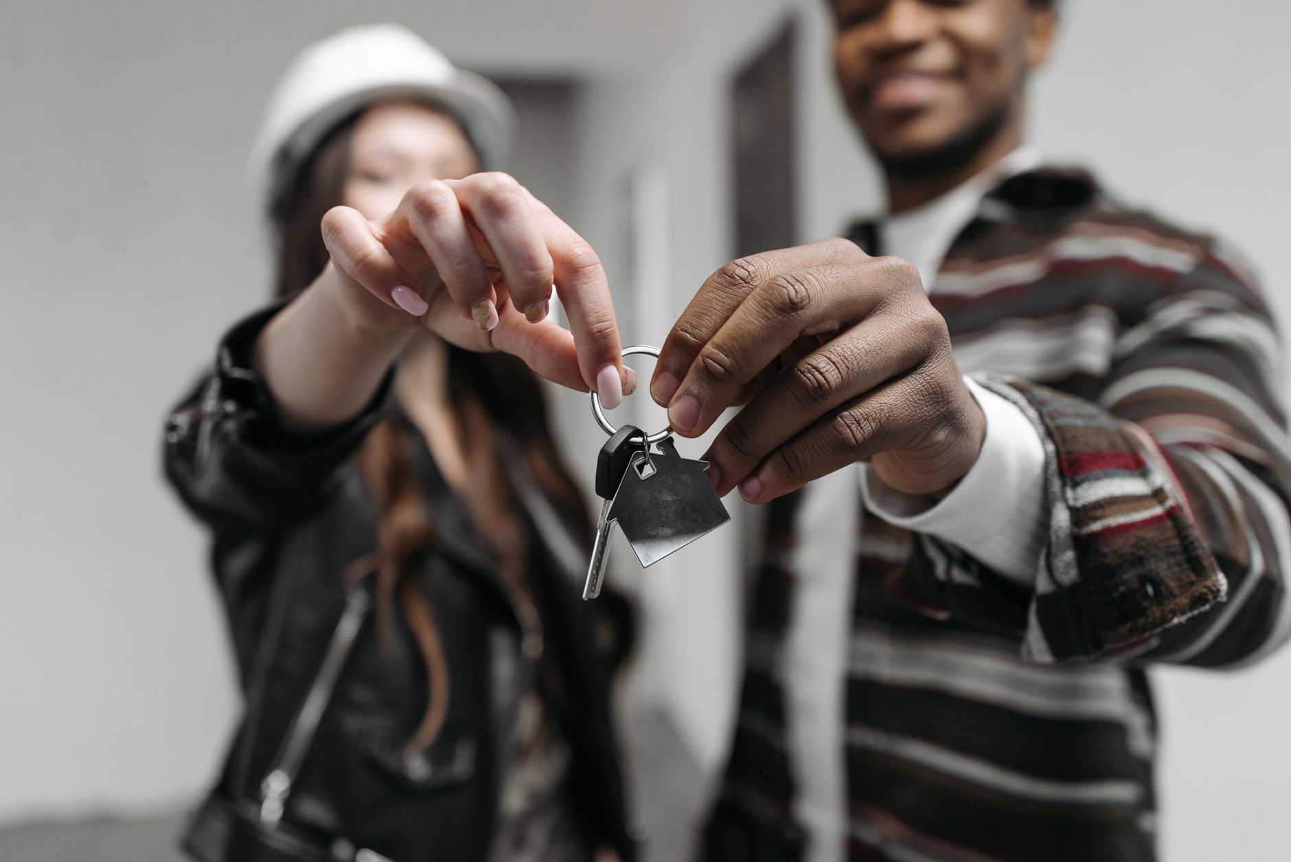 Deux personnes tendent des clés avec un porte-clés en forme de maison, dans une pièce blanche, le visage flou à l’arrière-plan.